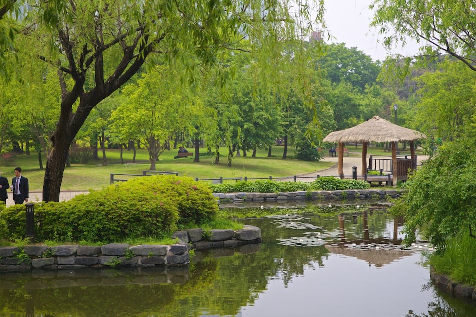 A serene park with a pond, lush greenery, and a thatched gazebo.