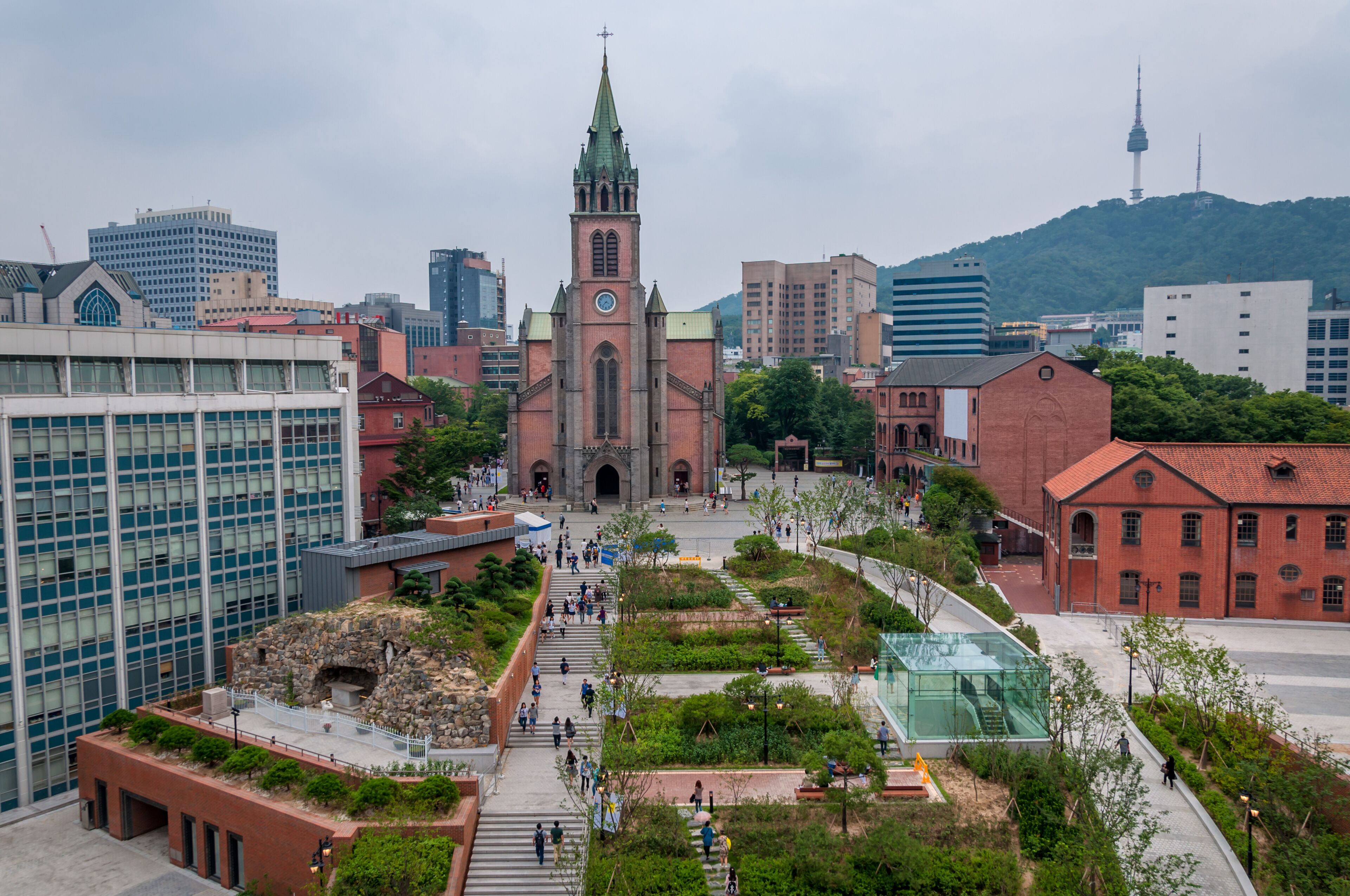 A church with a clock tower, surrounded by modern buildings and a landscaped area with pathways.