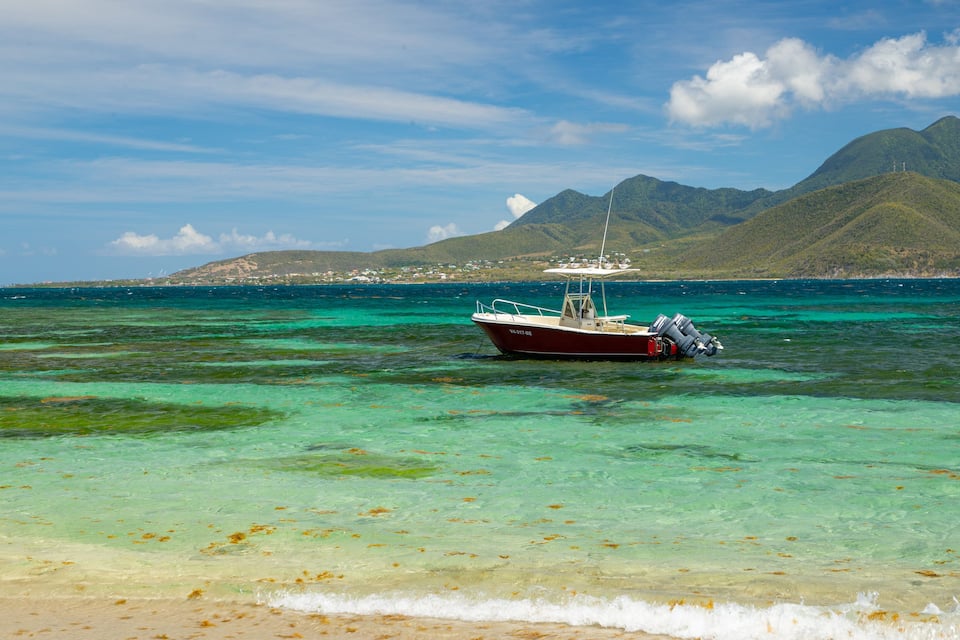 A boat anchored near a beach with clear turquoise water, sandy shore, and a mountainous backdrop.