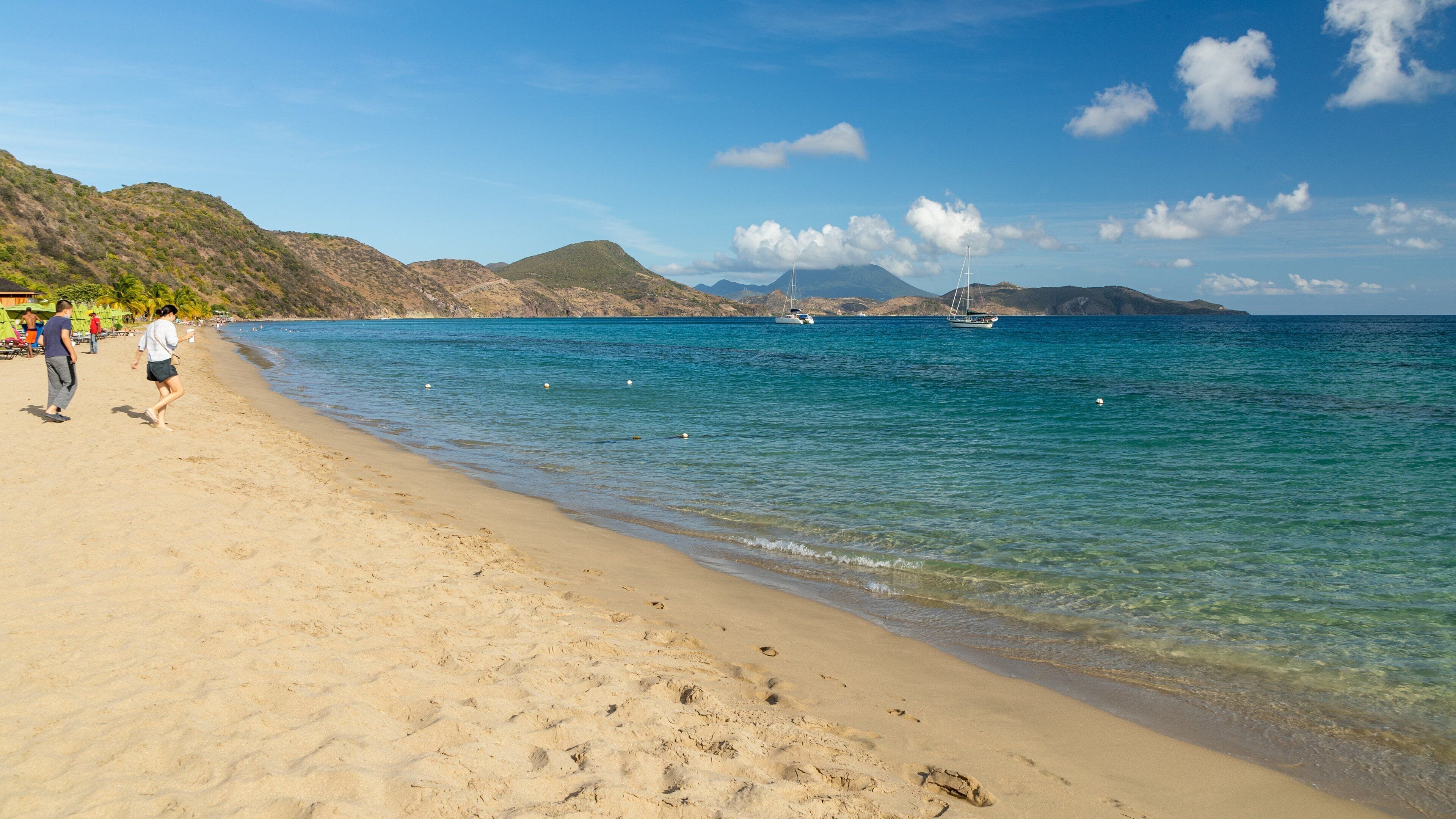 Une plage à l’eau turquoise et limpide, avec un rivage sablonneux et des bateaux visibles au loin.