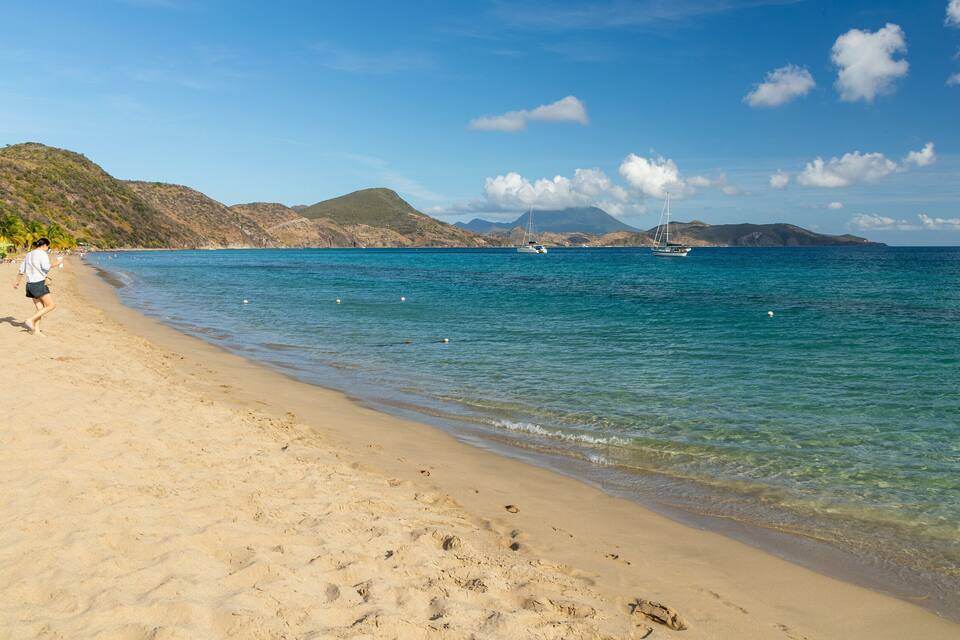 Une plage à l’eau turquoise et limpide, avec un rivage sablonneux et des bateaux visibles au loin.