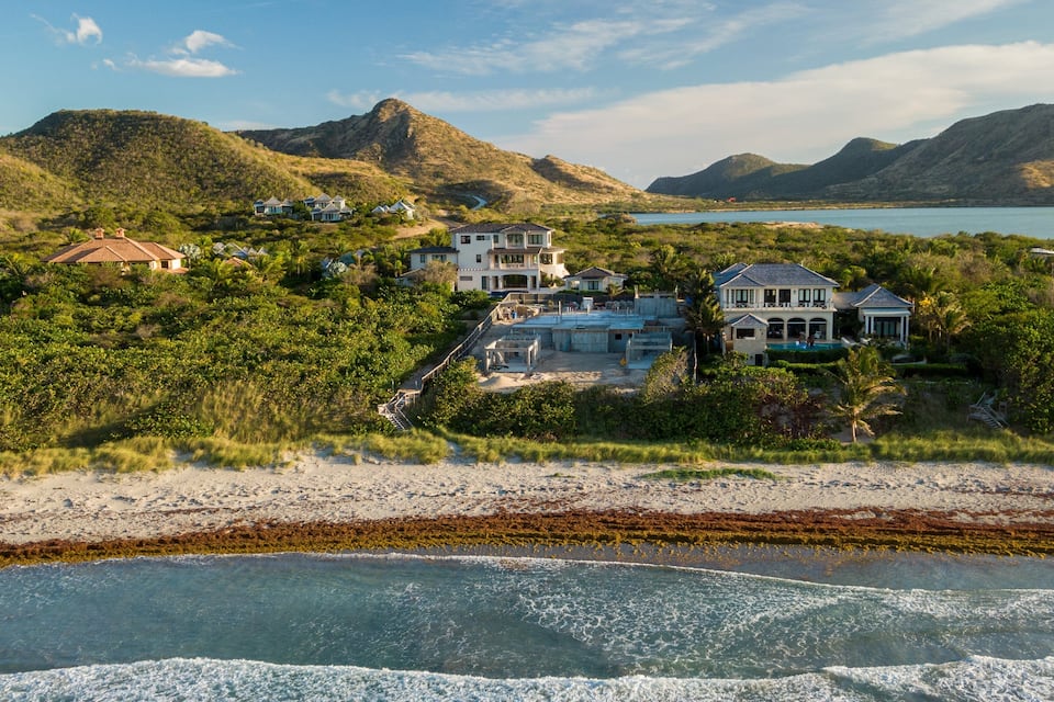 A coastal landscape with a beach, a large body of water, and a cluster of houses surrounded by greenery and hills.