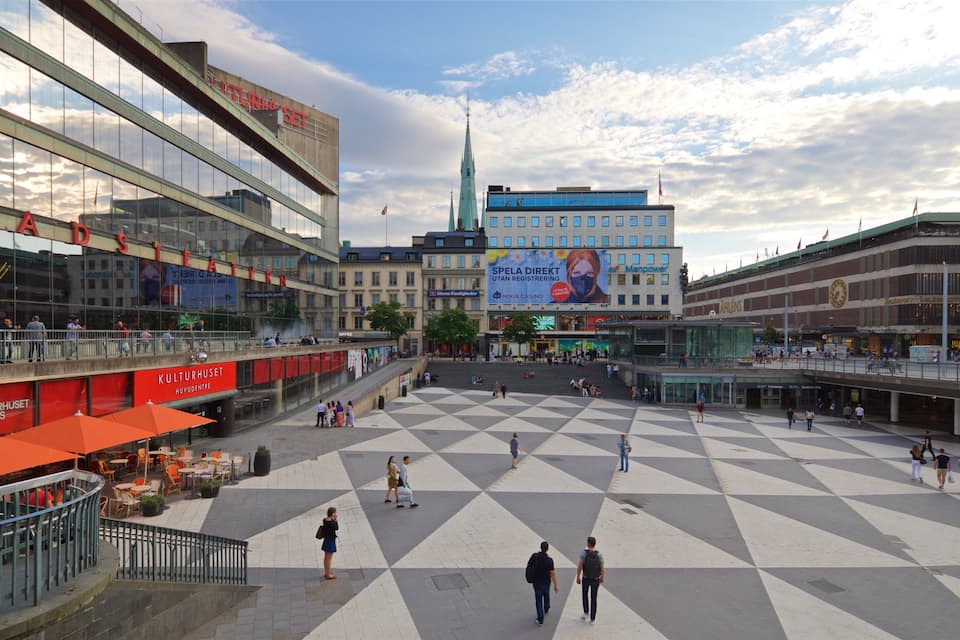 A public square with a large open space, surrounded by modern buildings, including a prominent building with a spire.