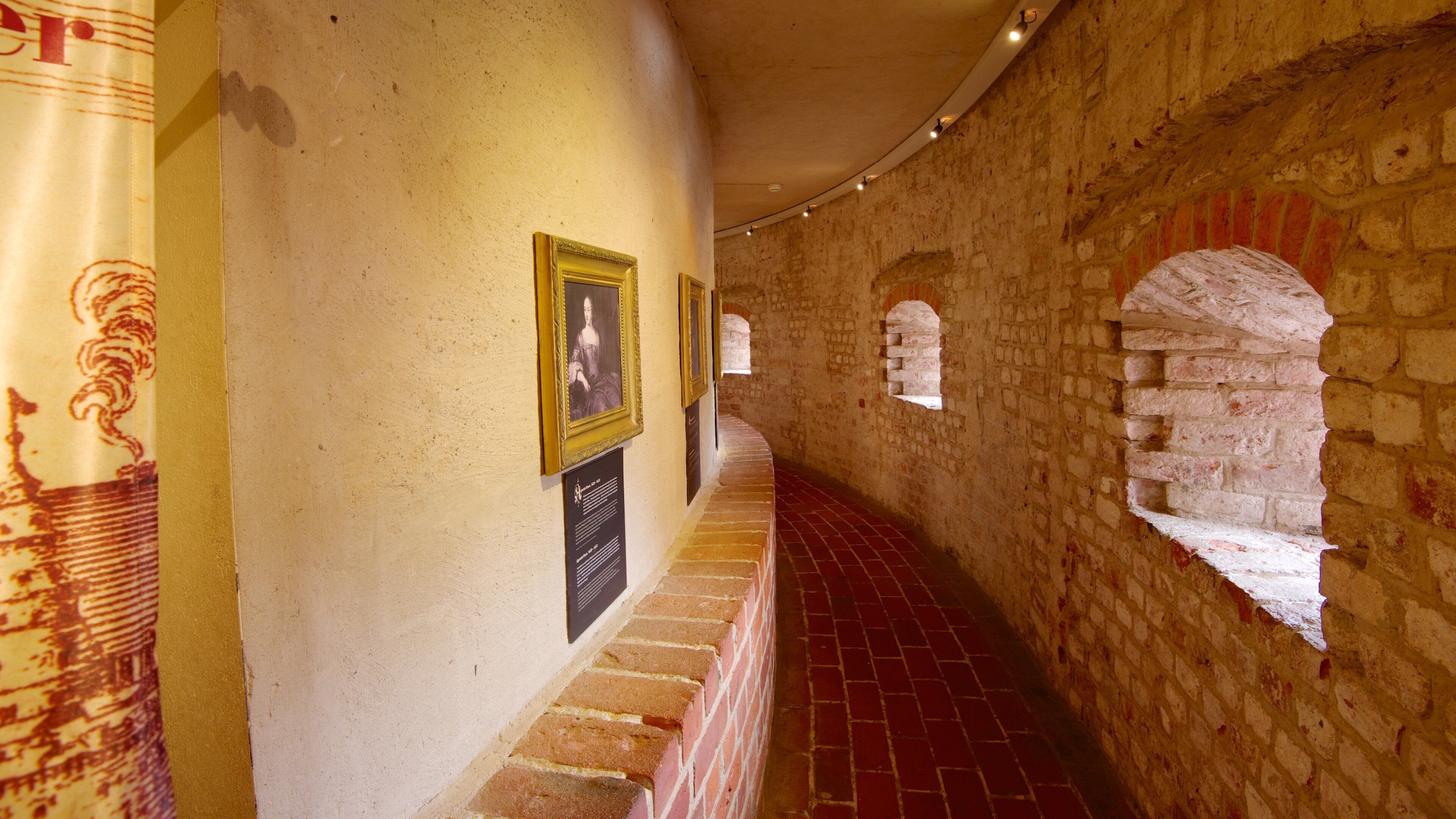 A corridor with stone walls, red brick flooring, and framed pictures on the wall.