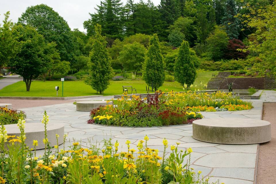 Um parque com um canteiro de flores circular, um caminho pavimentado e uma área gramada com árvores.