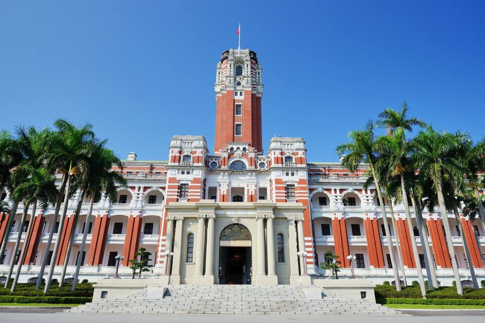 Un edificio grande e histórico con una torre central, fachada simétrica y palmeras al frente.