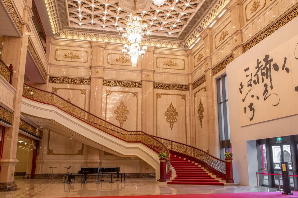 A grand interior with a red carpeted staircase, ornate gold detailing, and a large chandelier.