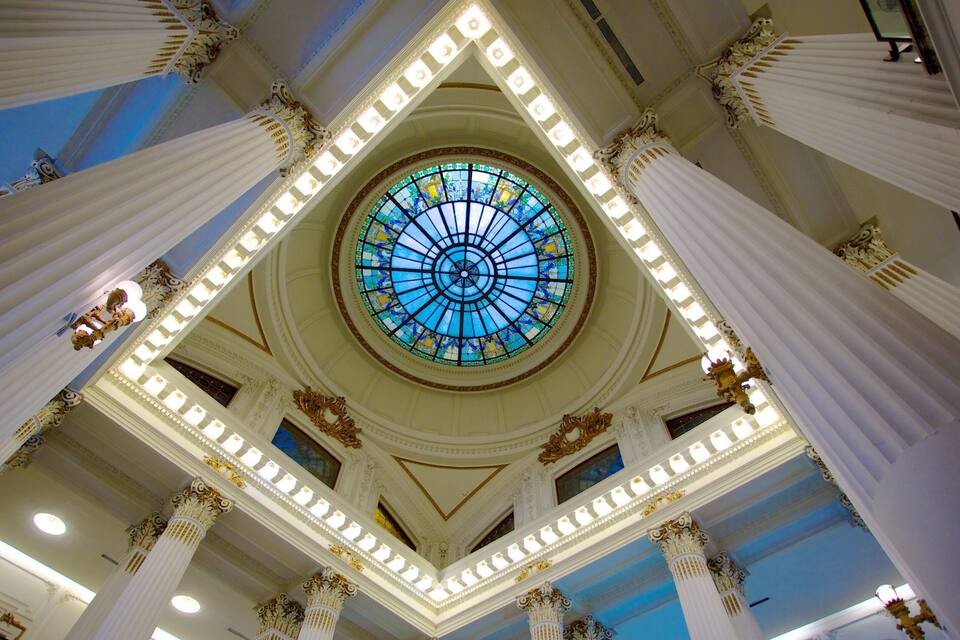 Un salón imponente con un amplio ventanal circular de vitrales, columnas ornamentadas y un techo alto.