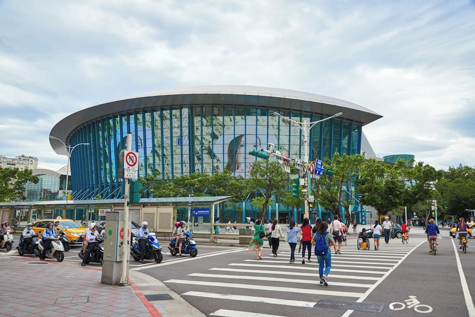 A modern building with a curved glass facade, a crosswalk, and people walking and riding bicycles.