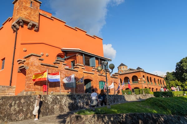 Un bâtiment historique à la façade en briques et doté d’une tour, entouré de drapeaux et de visiteurs.
