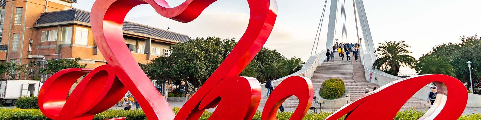 Una escultura roja con forma de corazón en un parque, con un puente moderno al fondo.