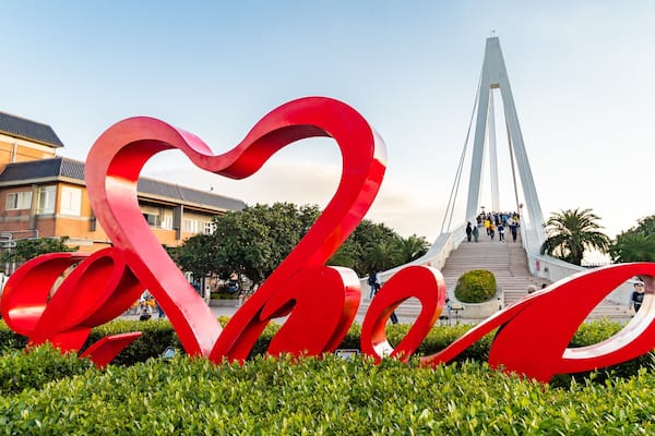 Eine herzförmige Skulptur in Rot in einem Park, im Hintergrund eine moderne Brücke.