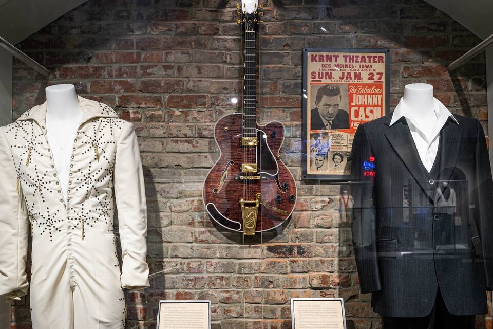 A display with mannequins in vintage clothing and a guitar, with a brick wall and a theater poster in the background.