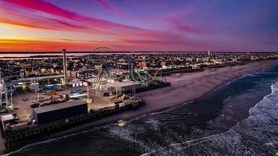 A coastal amusement park at sunset with a roller coaster, Ferris wheel, and beach.