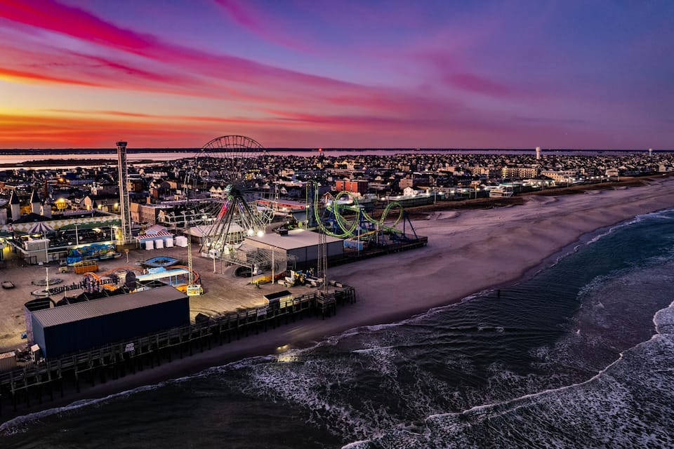A coastal amusement park at sunset with a roller coaster, Ferris wheel, and beach.