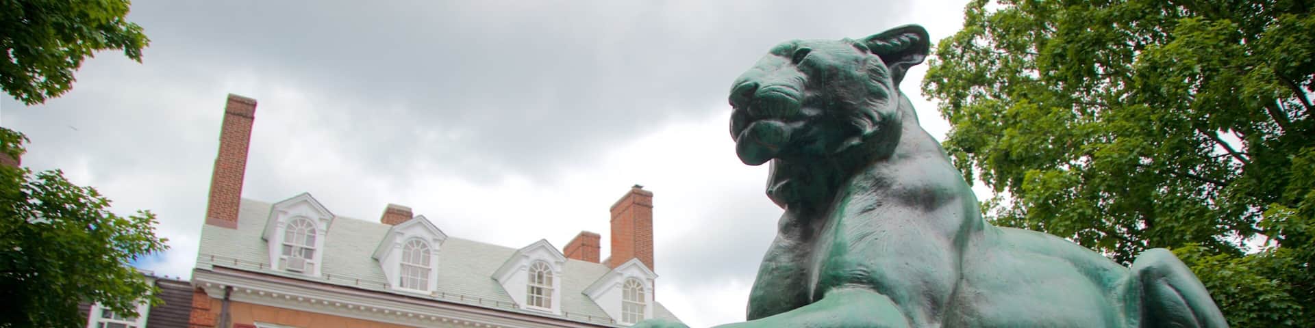 A bronze statue of a lion on a pedestal in front of a building with red shutters and a brick chimney.