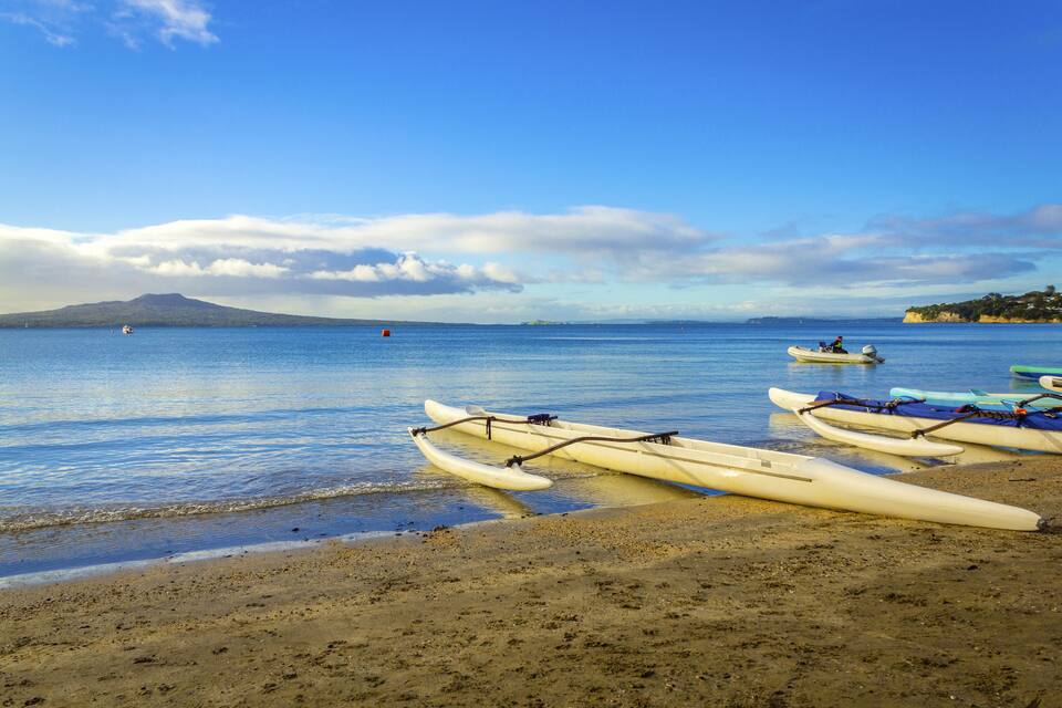 Una playa con varias kayaks alineadas en la orilla, una montaña distante y un cielo azul despejado.