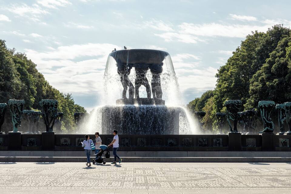 A large fountain with water jets, surrounded by trees and sculptures.