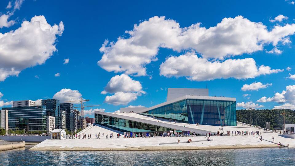 A waterfront cityscape with modern architecture, including a distinctive glass building and a large public square.