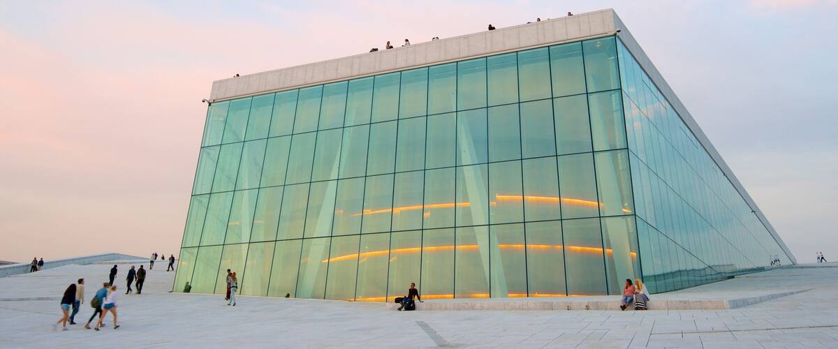 A modern glass building with a geometric design, surrounded by a spacious plaza with people walking and sitting.