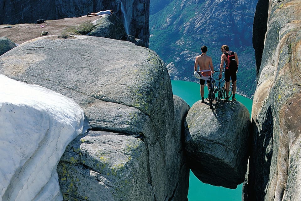 Duas pessoas em uma saliência rochosa observando um lago azul-turquesa com montanhas ao fundo.