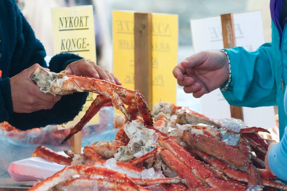 Uma pessoa segurando uma grande perna de caranguejo, com outras pernas de caranguejo e caixas com a etiqueta 'NYKÖKT KONGE-' ao fundo.