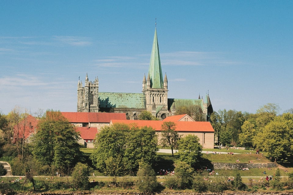 A historic church with a tall green spire, surrounded by trees and a park with people.