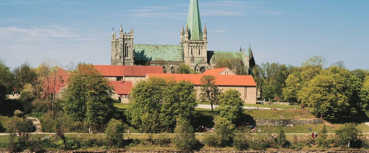 A historic church with a tall green spire, surrounded by trees and a park with people.