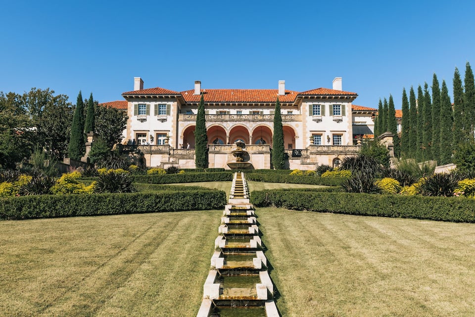 A grand estate with a symmetrical garden, a central fountain, and a clear blue sky.