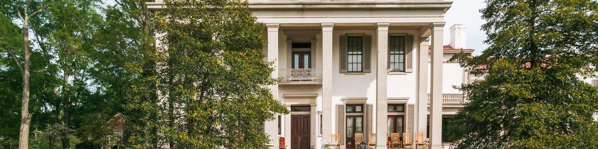 A large, stately building with columns and a balcony, surrounded by well-maintained lawns and trees.
