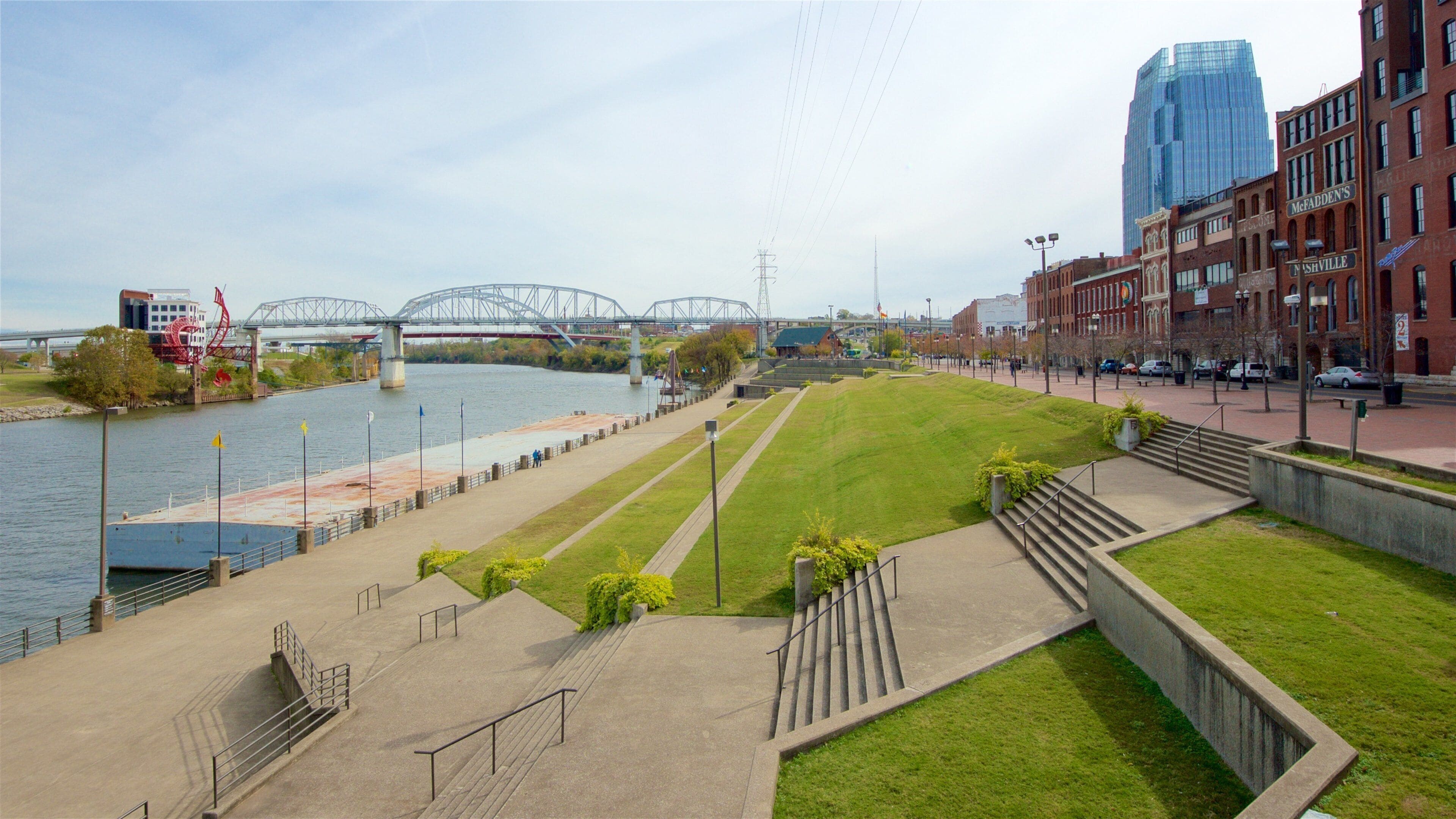 A riverside walkway with a bridge, grassy areas, and buildings.