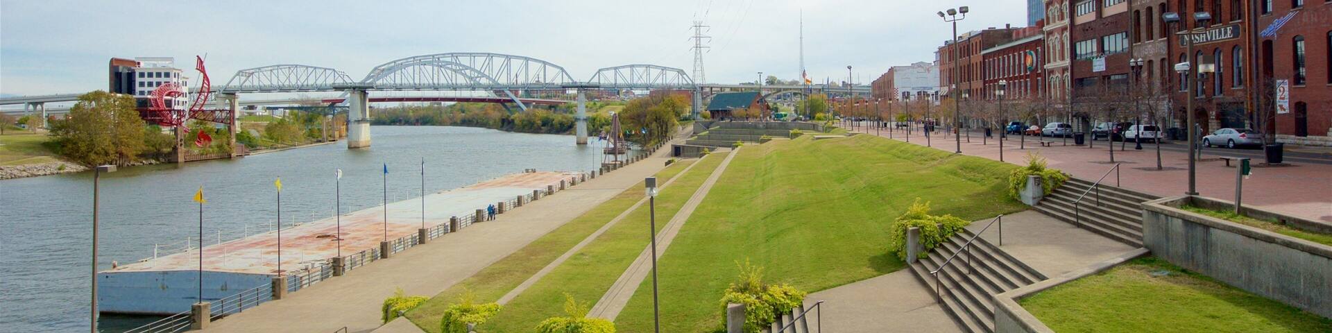 A riverside walkway with a bridge, grassy areas, and buildings.
