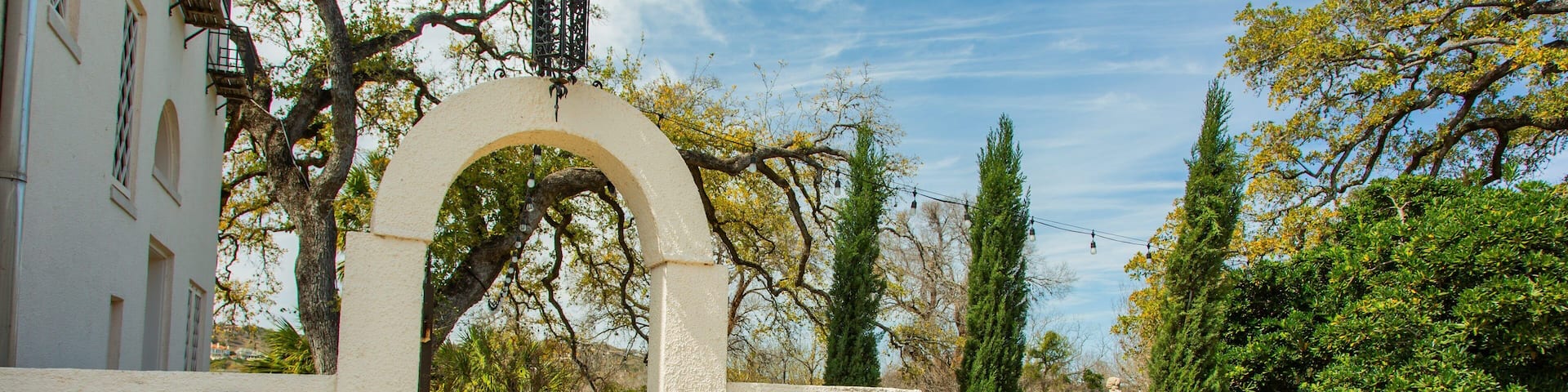 A stone archway leading to a courtyard with a statue, benches, and a building with a Texas flag.