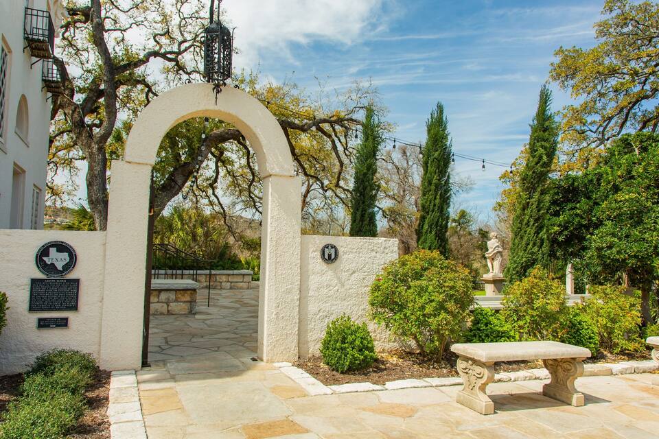 Ein steiner Torbogen führt in einen Innenhof mit einer Statue, Bänken und einem Gebäude mit einer Texas-Flagge.