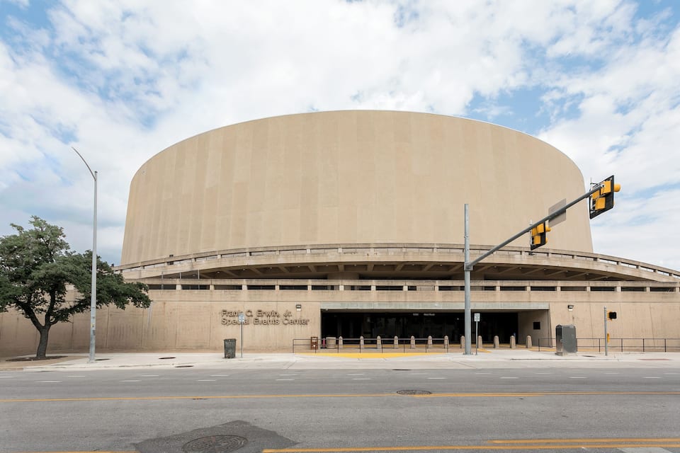 A large, beige, curved building with a flat roof, likely an event center, with a traffic light and a tree in the foreground.