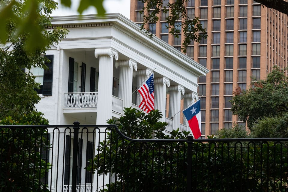 Um edifício branco com colunas, uma bandeira americana e a bandeira do estado do Texas.