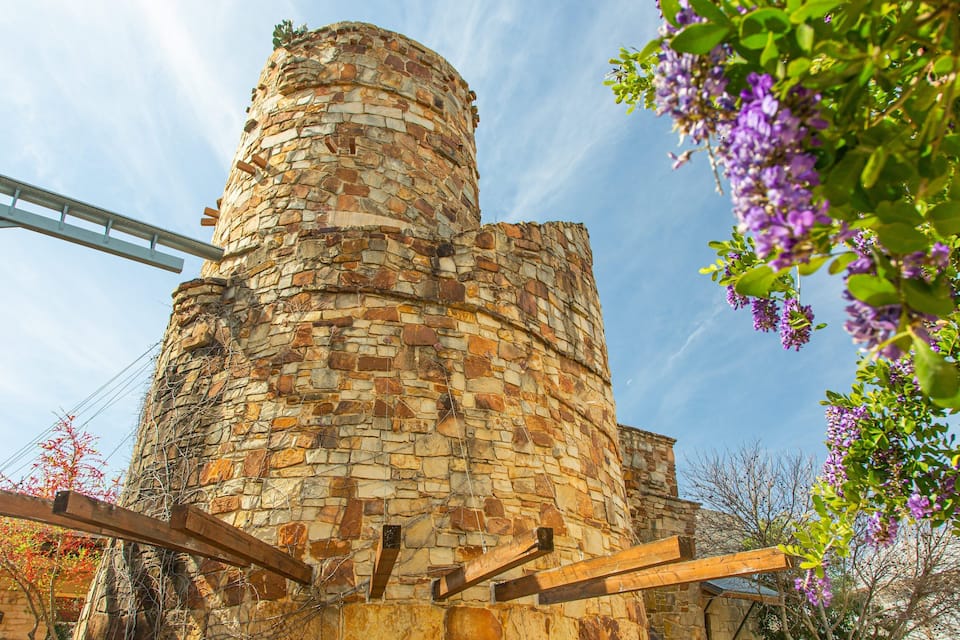 Una torre in pietra con una struttura metallica, circondata da alberi e fiori viola.