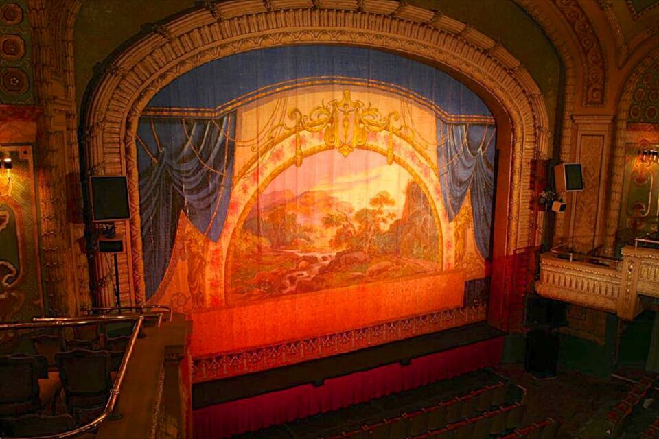 An ornate theater interior with a large mural, red curtains, and classical architecture.
