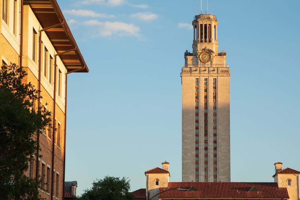 A tall clock tower with a clock face, surrounded by buildings with red-tiled roofs.