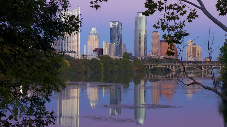 A city skyline reflected in a calm body of water, surrounded by trees and a bridge.