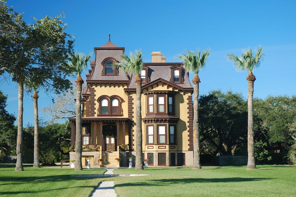 A large, historic house with a prominent tower and bay windows, surrounded by a well-maintained lawn and palm trees.