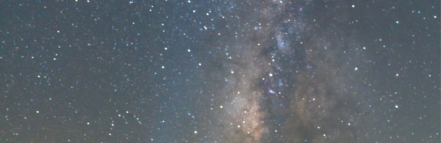 Céu noturno com estrelas e a Via Láctea visível sobre uma praia com um gazebo.