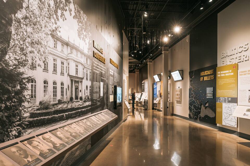 A museum corridor with historical exhibits, including a large wall with a building and text, and a display of photographs.