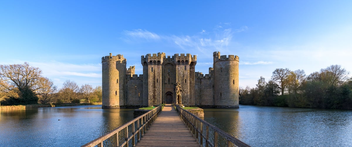A medieval castle with a wooden bridge leading to it, surrounded by water and trees.