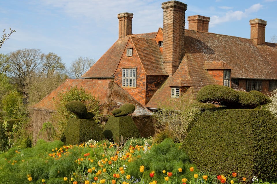 Une maison traditionnelle en briques, avec un toit en tuiles et plusieurs cheminées, entourée de jardins et de haies bien entretenus.