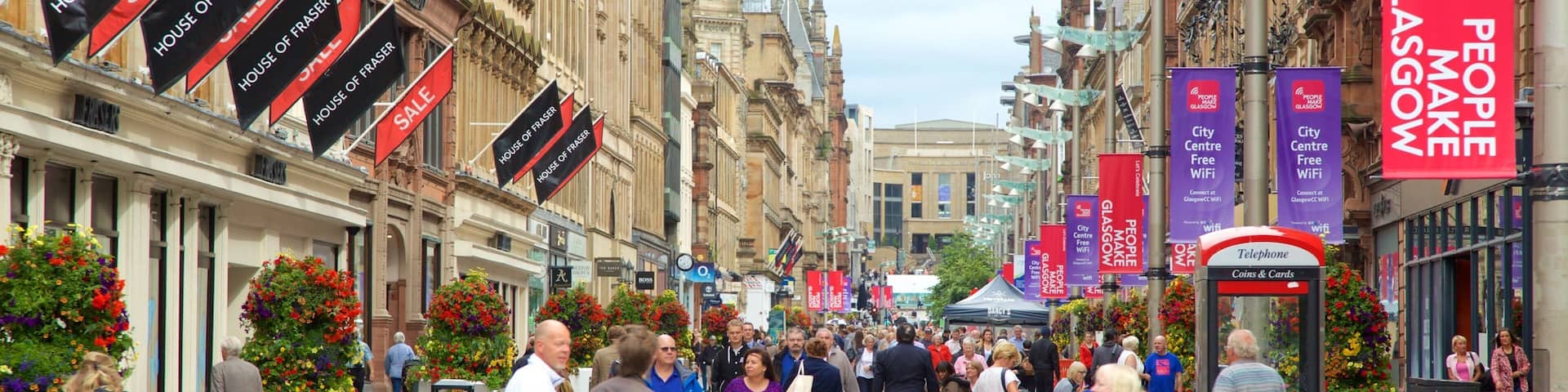 A busy street in Glasgow with people walking, a bus stop, and various banners advertising events and sales.