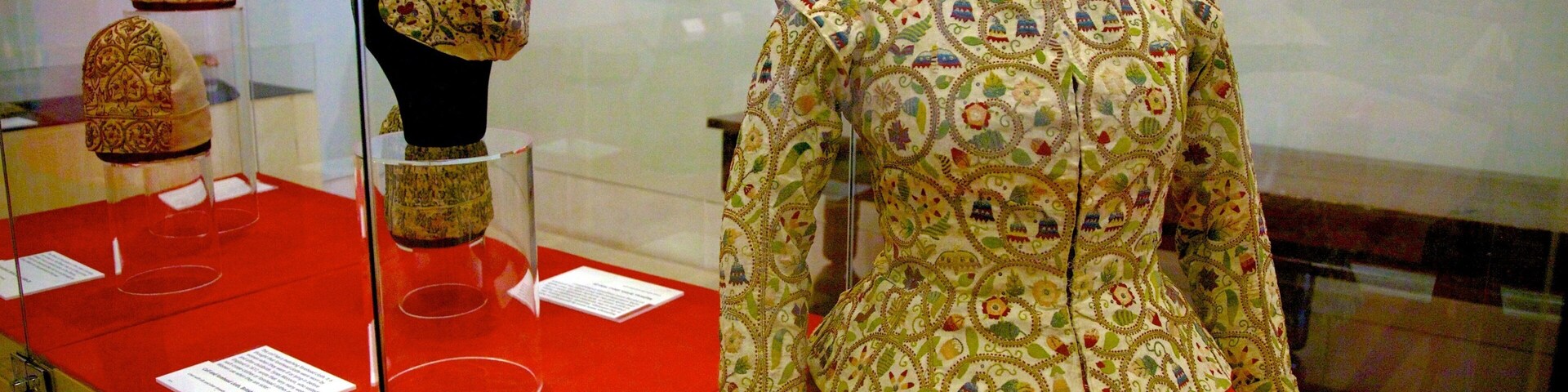Mannequin em museu exibindo uma peça de vestuário ricamente bordada, com saia vermelha.