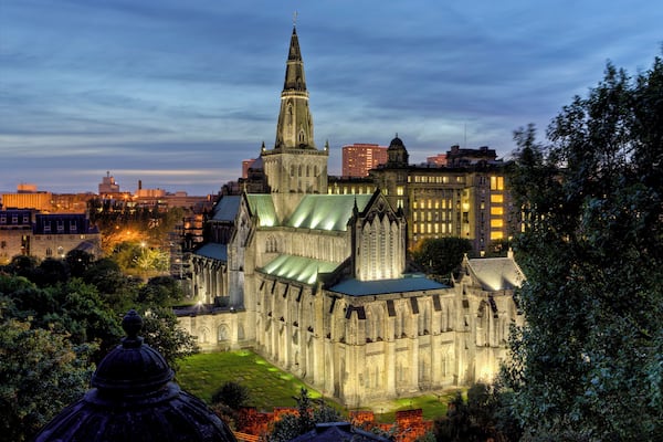 A historic cathedral with a tall spire, surrounded by modern buildings and lush greenery.