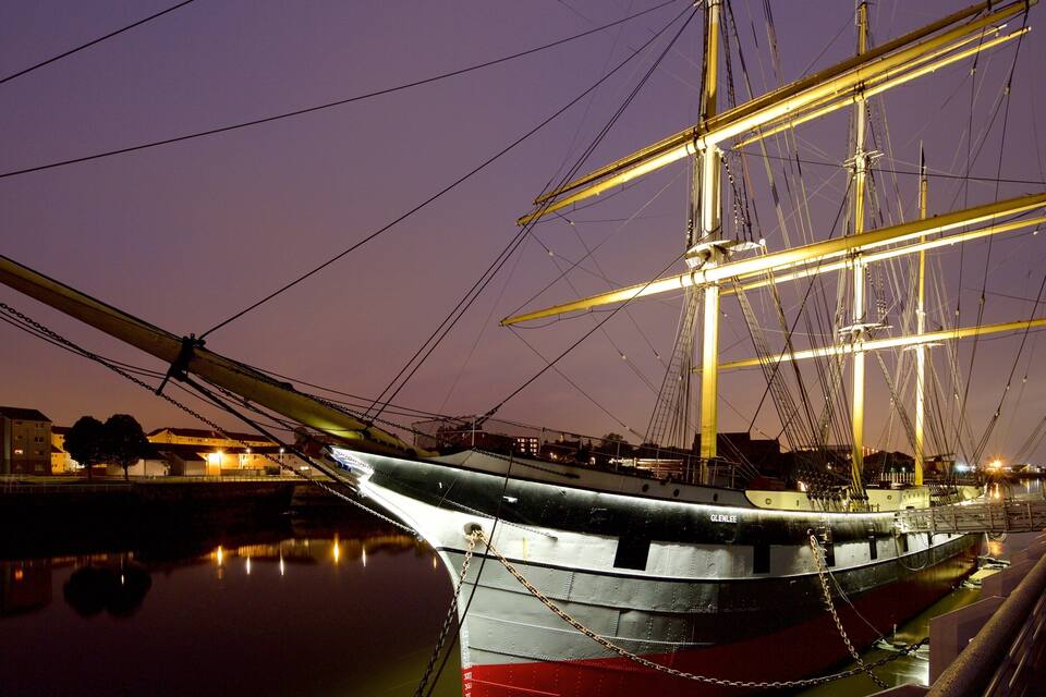 Un navire à voile historique est amarré dans un port, avec des bâtiments en arrière-plan.