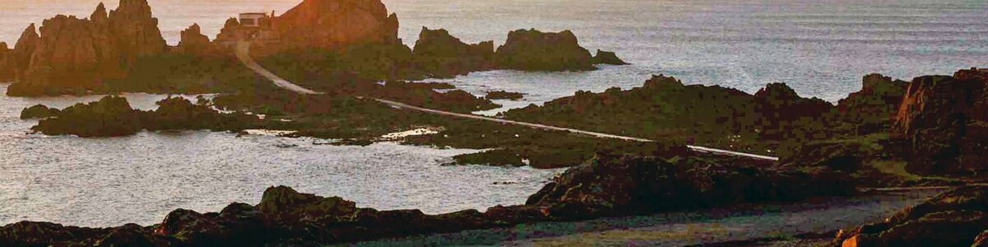 A lighthouse on a rocky outcrop with a road leading to it, surrounded by calm sea and clear sky.